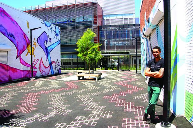 ➥➥ Foto por Yadira Betances. Marquis Victor, director del programa Elevated Thought posa en frente del mural pintado por tres artistas del programa en el callejón detrás del Dr. Ibrahim El-Hefni Allied Health and Technological Center de Northern Essex Community College.
